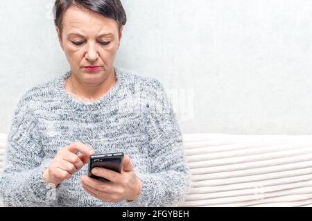 Porträt einer älteren Frau Aussehen Telefon, während auf dem Sofa sitzen Stockfoto