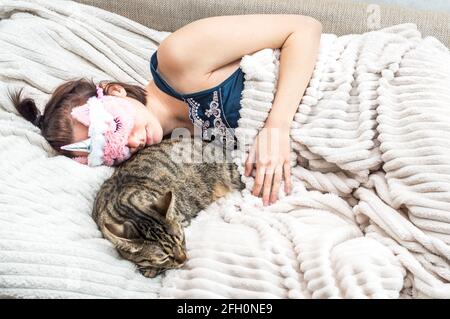 Porträt einer jungen Frau mit einer Schlafmaske im Gesicht und einem Schlafanzüge, der ihre Katze umarmt. Konzept von Ruhe und Schlaf. Stockfoto