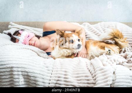Porträt einer jungen Frau mit Schlafmaske und Schlafanzügen im Bett. In der Nähe ist ein roter Hund Stockfoto