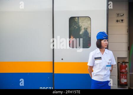 Almaty, Kasachstan - 06. Juni 2012: Porträt einer jungen asiatischen Zugwirtin in Garisson-Mütze. Talgo-Tulpar-Schnellzug im Hintergrund. Stockfoto