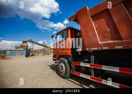 Gewinnung von Golderz. Bergbau- und Verarbeitungsanlage.Orange LKW Transport von Golderz zu gelben Gesteinszerdrückungsmaschine. Blauer Himmel, Wolken. Stockfoto