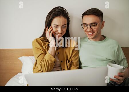 Schönes Paar, das sich mit romantischen Gefühlen einen Film ansieht und mit Kaffee auf dem Bett sitzt. Glückliche Familie Umzug in neue Wohnung Stockfoto