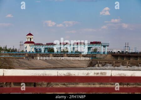 Damm auf dem Fluss Shardara. Hebevorrichtung der Wasserblenden. Alte sowjetische Brücke und Hochhaus. Kyzylorda, Kasachstan Stockfoto