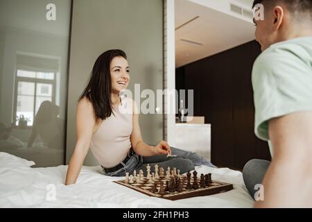 Glückliches junges Paar verbringt ihre Freizeit mit Schachspielen, auf dem Bett. Junge Familie zieht in eine neue Wohnung Stockfoto