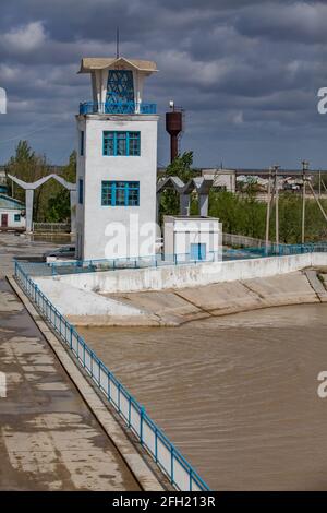 Sowjetische Vintage Turm Gebäude auf Shardara Fluss Damm und Brücke.verrostet Wasserspeicher Turm rechts.Gelbes Wasser.Kazaly Stadt, Kyzylorda Region, Kasachstan Stockfoto