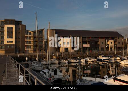 Boote, die in Victoria Dock, Caernarfon, Wales bei Abendsonne festgemacht wurden. Apartments und Restaurants im Hintergrund Stockfoto