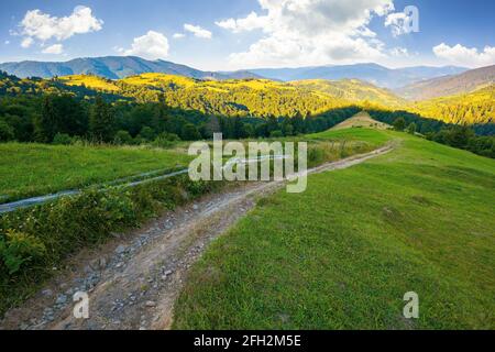 Ländliche Landschaft in den Bergen bei Sommersonnenaufgang. Landstraße durch grasbewachsene Weide, die hinunter in das ferne Tal. Wolken auf dem blauen Himmel abov