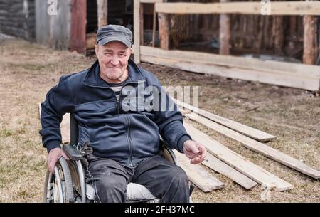 Porträt eines glücklichen älteren Mannes in legerer Kleidung in einem Rollstuhl im Hof seines Hauses, der lächelt und die Kamera anschaut. Eine Zigarette in der Hand. Stockfoto