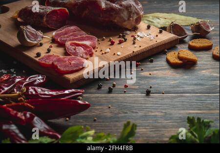 In Scheiben geschnitten, ruckartig. Wurststab auf das Brett geschnitten. Knoblauch, rote Paprika und grüne Petersilie in der Nähe der Aufschnitte. Stockfoto