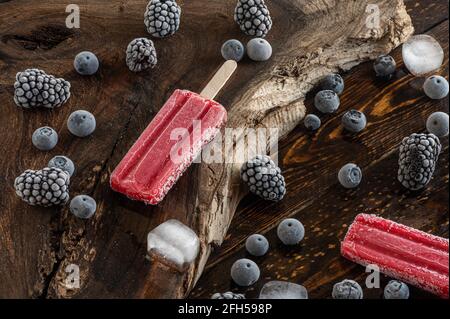 Gefrorene Brombeeren, Heidelbeeren, Eislollies und Eiswürfel über einem Holzbrett Stockfoto