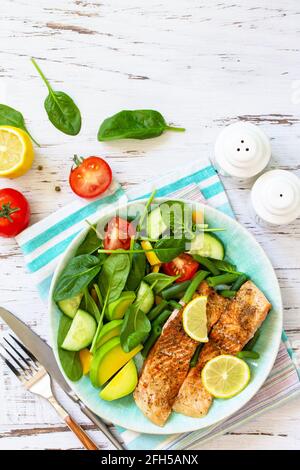 Gegrilltes Lachsfilet aus nächster Nähe mit Spinat, Gurke, Tomaten und Avocado-Salat auf einem weißen Holztisch. Draufsicht, flach liegend. Speicherplatz kopieren. Stockfoto