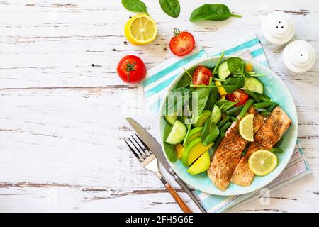 Gegrilltes Lachsfilet aus nächster Nähe mit Spinat, Gurke, Tomaten und Avocado-Salat auf einem weißen Holztisch. Draufsicht, flach liegend. Speicherplatz kopieren. Stockfoto