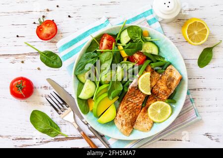 Gegrilltes Lachsfilet aus nächster Nähe mit Spinat, Gurke, Tomaten und Avocado-Salat auf einem weißen Holztisch. Draufsicht, flach liegend. Stockfoto