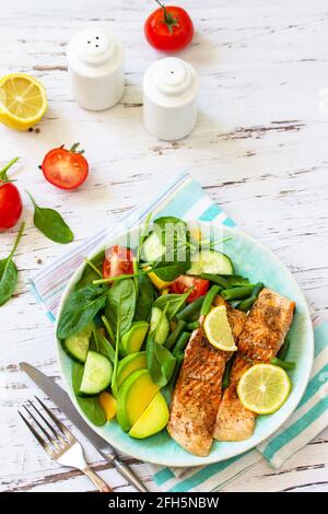 Gegrilltes Lachsfilet aus nächster Nähe mit Spinat, Gurke, Tomaten und Avocado-Salat auf einem weißen Holztisch. Speicherplatz kopieren. Stockfoto