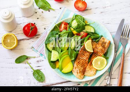 Gegrilltes Lachsfilet mit Spinat, Gurke, Tomaten und Avocado-Salat auf einem weißen Holztisch. Stockfoto
