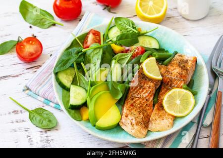 Gegrilltes Lachsfilet aus nächster Nähe mit Spinat, Gurke, Tomaten und Avocado-Salat auf einem weißen Holztisch. Stockfoto