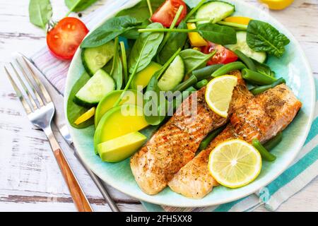 Gegrilltes Lachsfilet aus nächster Nähe mit Spinat, Gurke, Tomaten und Avocado-Salat auf einem weißen Holztisch. Stockfoto