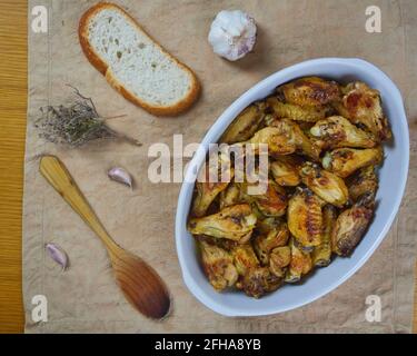 Knoblauch-Hühnerflügel (Pollo al ajillo). Spanisches Tapas-Gericht in einer Keramikplatte, serviert mit Knoblauchhandschuhen, knusprigem Brot, Blumen und einem Holz Stockfoto