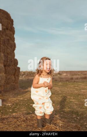 Unbeschwertes Kind in Overalls, das auf Strohballen bei Sonnenschein sitzt Tag auf dem Land Stockfoto