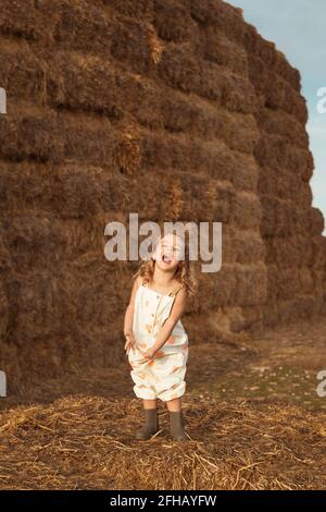 Unbeschwertes Kind in Overalls, das auf Strohballen bei Sonnenschein sitzt Tag auf dem Land Stockfoto