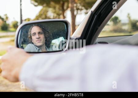 Lächelnder junger männlicher Fahrer, der sich im Rückspiegel der Seite widerspiegelte Des Autos geparkt auf grüner Wiese während der Sommerreise durch Auf dem Land Stockfoto