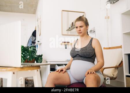 Junge, werdende Frau, die auf dem Gymnastikball sitzt und in einem Laptop surft Im Wohnzimmer Stockfoto