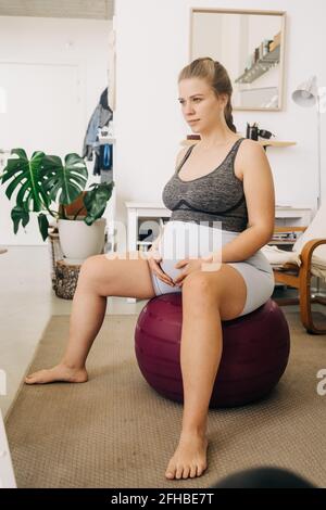 Junge, werdende Frau streichelte den Bauch, während sie auf dem Gymnastikball saß Und im Wohnzimmer wegschauen Stockfoto