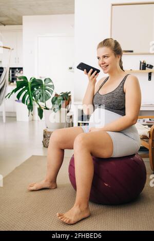 Junge Inhalte erwartungsvolle weibliche sitzen auf Fitness-Ball während des Sendens Sprachnachricht auf dem Handy und der Blick weg zu Hause Stockfoto