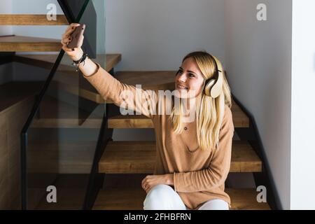 Positive Frau, die Musik über Kopfhörer hört und sich selbst nimmt Aufgenommen auf dem Smartphone, während Sie zu Hause auf einer Holztreppe sitzen Und am Wochenende unterhaltsam Stockfoto