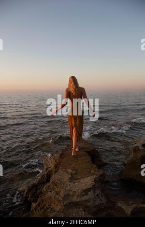 Rückansicht einer jungen Frau, die bei Sonnenuntergang an der Küste gegen das blau winkende Meer läuft Stockfoto