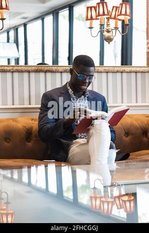 Fokussierter afroamerikanischer Unternehmer in eleganten Kleidern, auf dem er sitzt Sofa im Luxuscafe und Lesebuch Stockfoto