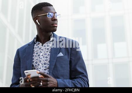 Junger, gut gekleideter afroamerikanischer Geschäftsmann mit Brille und kabellosem Internetzugang Ohrstöpsel mit Mobiltelefon, das wegschaut Stockfoto