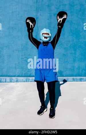 In voller Länge lächelnde reife Frau in Sportswear und Boxhandschuhen Stehen mit Helm gegen blaue Wand mit erhobenen Armen Stockfoto