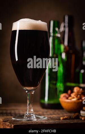 Glas dunkles Bier mit Schaumstoffkopf auf dunklem Holz Hintergrund mit leeren Flaschen und Biersnacks Stockfoto