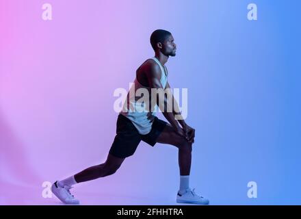 Seitenansicht des fitgen schwarzen Mannes in Sportswear, der Ausfallschritte im neonfarbenen, langen Porträt macht. Gesundes Wohnkonzept Stockfoto