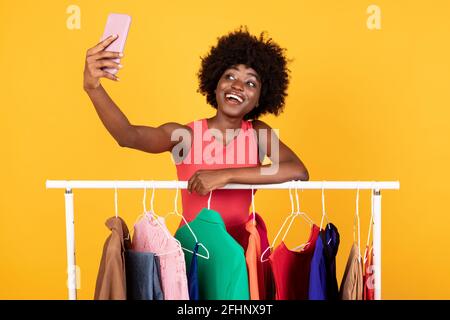 Black Lady Making Selfie Auf Dem Smartphone In Der Nähe Von Clothing Rail, Studio Stockfoto