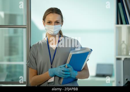 Porträt eines attraktiven Laborforschers in Gesichtsmaske und Handschuhen Mit Abzeichen am Hals, der an der Tür gelehnt ist und einen Ordner hält Mit Papieren Stockfoto