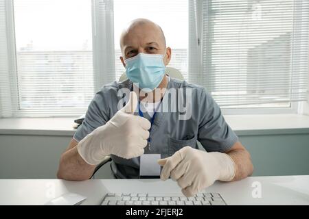 Porträt eines zufriedenen kahlen Arztes in Gesichtsmaske und Latex Handschuhe sitzen am Tisch und zeigen den Daumen nach oben Stockfoto