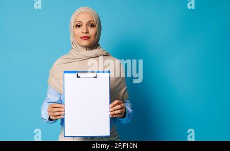 Schöne arabische Frau, die einen beigen Hijab trägt und eine Zwischenablage mit weißem, leerem Blatt Papier hält, isoliert auf blauem Hintergrund Stockfoto