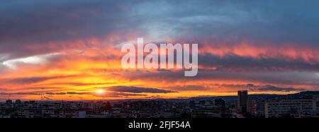 Dramatisches Sonnenuntergangsfeuer am Himmel über einer kleinen Stadt. Die Abendsonne ist tief am Horizont. Panoramalandschaft mit urbaner Skyline. Lebendiger Himmel bei Sonnenuntergang Stockfoto