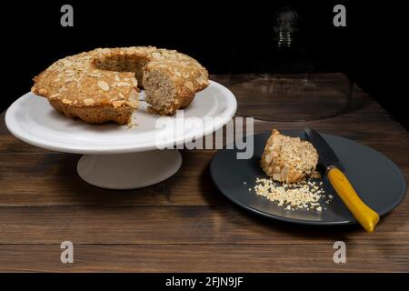 Eine Scheibe Mandeldonut mit Buchweizen auf einem dunklen Teller neben einem weißen Kuchenständer mit dem Rest Des Kuchens oben Stockfoto
