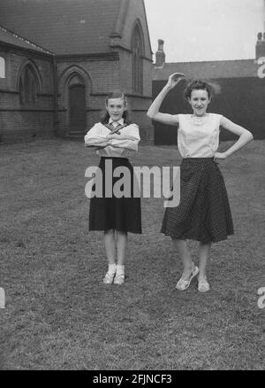 1956, historisch, draußen auf dem Gelände der Kirche, zeigen zwei Teenager-Mädchen Tanzbewegungen, die sie am 1. Mai-Karneval, LEDs, England, Großbritannien, durchführen werden. Ein uraltes Fest, das die Ankunft des Frühlings feiert, beinhaltete die Krönung einer May Queen und das Tanzen um einen Maypole, Aktivitäten, die seit Jahrhunderten in England stattfinden. Ausgewählt aus den Mädchen der Gegend, die Mai-Königin würde die Prozession der Wagen und Tanz zu starten. Im industrialisierten Norden Englands führten die Kirchensonntagsschulen oft die Organisation des Karnevals an. Stockfoto