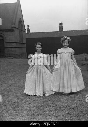 1956, historisch, draußen auf dem Gelände der Kirche, zeigen zwei junge Mädchen ihre hübschen Kleider für den 1. Mai-Karneval, wo sie die May Queen des Jahres in ihren Pflichten unterstützen werden, Leeds, England, Großbritannien. Ein uraltes Fest, das die Ankunft des Frühlings feiert, beinhaltete die Krönung einer May Queen und das Tanzen um einen Maypole, Aktivitäten, die seit Jahrhunderten in England stattfinden. Ausgewählt aus den Mädchen der Gegend, die Mai-Königin würde die Prozession der Wagen und Tanz zu starten. Im industrialisierten Norden Englands führten oft die kirchlichen Sonntagsschulen die Organisation an. Stockfoto