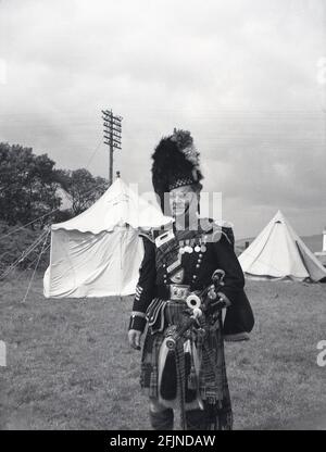 1956, historisch, ein schottischer Pfeifer, der in seiner Uniform und mit Dudelsack für ein Foto bei den Greenock Highland Games, Schottland, steht. Erstmals in den 1820er Jahren in Invergarry dokumentiert, zeigten die Einheimischen bei den Spielen ihre Stärke und Agilität bei verschiedenen Aktivitäten wie dem Werfen des Baumstamms und dem Hammerwurf. Sie beinhalteten auch schottischen Tanz und Piping. Der Besuch von Königin Victoria auf dem Braemer-Treffen im Jahr 1848 begann die Royal Association und sah sie in der Popularität zu erhöhen. In den Sommermonaten finden in ganz Schottland verschiedene Veranstaltungen statt, die als Highland Games oder einfach nur Versammlungen bekannt sind. Stockfoto