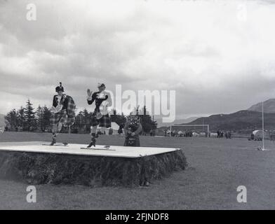 1956, ein Mann und eine Frau, die auf einer hölzernen Plattform traditionell schottisch tanzen oder auf einem Feld bei einem Hochlandspiel in Greenock, Schottland, Großbritannien, spielen. Erstmals in den 1820er Jahren in Invergarry dokumentiert, zeigten die Einheimischen bei verschiedenen Aktivitäten wie dem Hammerwerfen ihre Stärke und Agilität. Sie beinhalteten auch schottische Tänzen und Piping. Der Besuch von Königin Victoria auf dem Braemer-Treffen im Jahr 1848 begann die Royal Association und sah sie in der Popularität zu erhöhen. In den Sommermonaten finden in ganz Schottland verschiedene Veranstaltungen statt, die als Highland Games oder einfach nur Versammlungen bekannt sind. Stockfoto