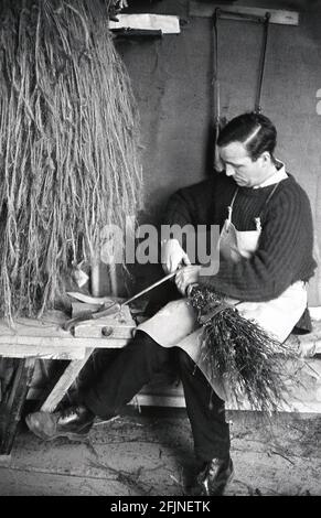 1950, historisch, ein männlicher Handwerker in seiner Werkstatt, die einen Besen herstellte, England, Großbritannien. Ein altes Handwerk, die Herstellung von Besen, auch bekannt als "Besoms" oder "Besom making", besteht aus einem Paket von Baumzweigen, die sicher um einen Stock gebunden sind. Traditionell wurden Besen aus Zweigen der Birke hergestellt, und ein Dorfbesbeer und ein Kupferarbeiter produzierten sie beide. Heute ist dieser hochspezialisierte Handwerksbetrieb, wie viele alte ländliche Handwerksbetriebe, fast vollständig ausgestorben, und es gibt heute weniger als fünf Besen-Hersteller im Vereinigten Königreich, die Besen auf einer Kommerzbasis herstellen. Stockfoto