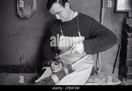 1950, historisch, ein männlicher Handwerker in seiner Werkstatt, die einen Besen herstellte, England, Großbritannien. Ein altes Handwerk, die Herstellung von Besen, auch bekannt als "Besoms" oder "Besom making", besteht aus einem Paket von Baumzweigen, die sicher um einen Stock gebunden sind. Traditionell wurden Besen aus Zweigen der Birke hergestellt, und ein Dorfbesbeer und ein Kupferarbeiter produzierten sie beide. Heute ist dieser hochspezialisierte Handwerksbetrieb, wie viele alte ländliche Handwerksbetriebe, fast vollständig ausgestorben, und es gibt heute weniger als fünf Besen-Hersteller im Vereinigten Königreich, die Besen auf einer Kommerzbasis herstellen. Stockfoto
