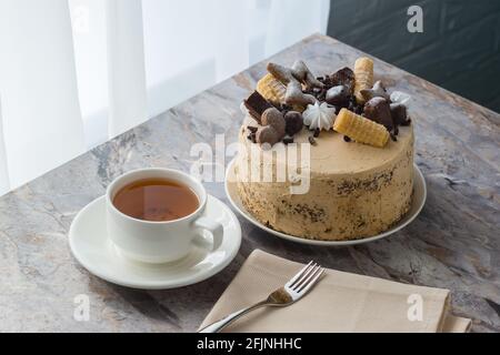 Eine Tasse Tee auf einer Untertasse und ein Biskuitkuchen mit Buttercreme auf der Marmorplatte. Der Kuchen ist mit Schokolade und Keksen dekoriert Stockfoto