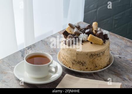 Eine Tasse Tee auf einer Untertasse und ein Biskuitkuchen mit Buttercreme auf der Marmorplatte in der Nähe des Fensters. Der Kuchen ist mit Schokolade und Keksen dekoriert Stockfoto