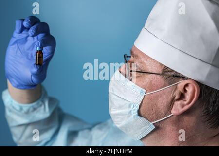Ein älterer Arzt in medizinischer Kleidung: Mütze, Handschuhe, Morgenmantel und Maske, Brille. Untersucht eine Flasche Medizin oder Impfstoff Stockfoto